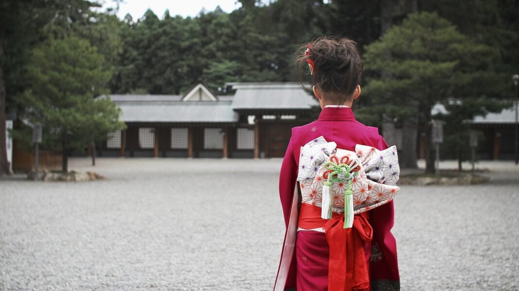 A young girl in a red kimono with an obi bow standing in a shrine courtyard for Shichi-Go-San